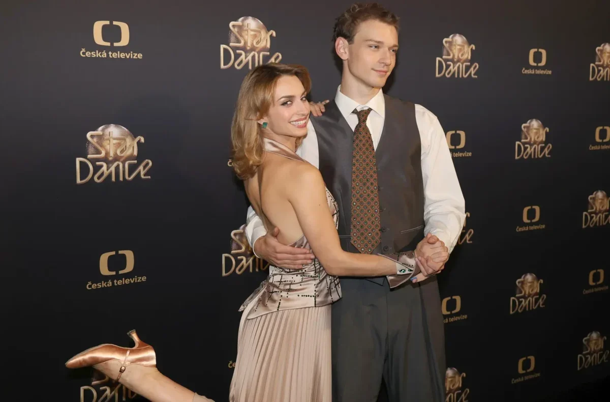 Smiling couple posing and dancing on a black backdrop with Star Dance and Česká televize logos; woman in a champagne dress and man in a gray vest and tie present on a red carpet backdrop.