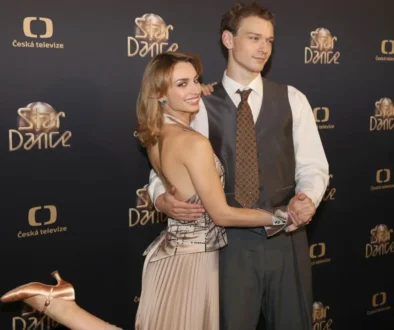 Smiling couple posing and dancing on a black backdrop with Star Dance and Česká televize logos; woman in a champagne dress and man in a gray vest and tie present on a red carpet backdrop.