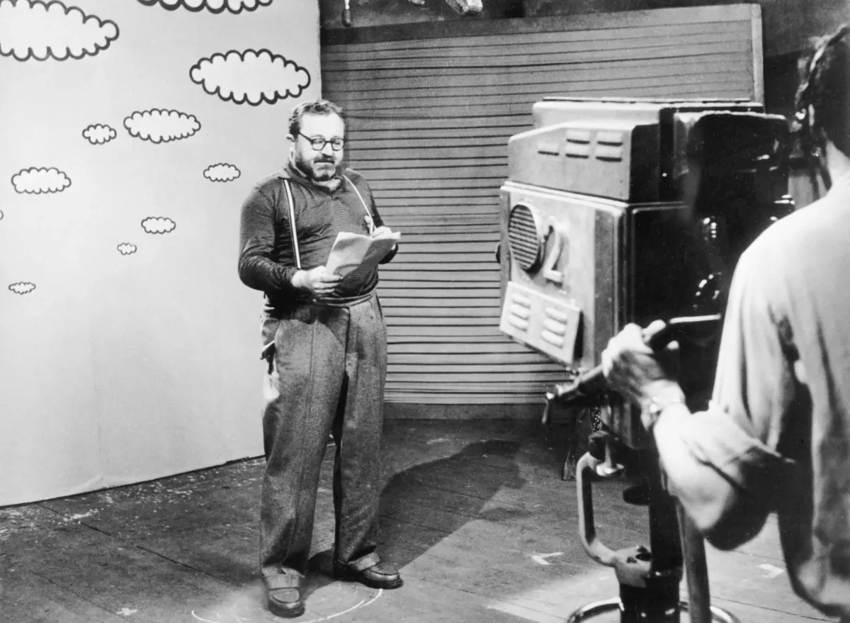 Man with glasses and suspenders reads a paper on a film set with a cartoon cloud backdrop and a large vintage camera nearby