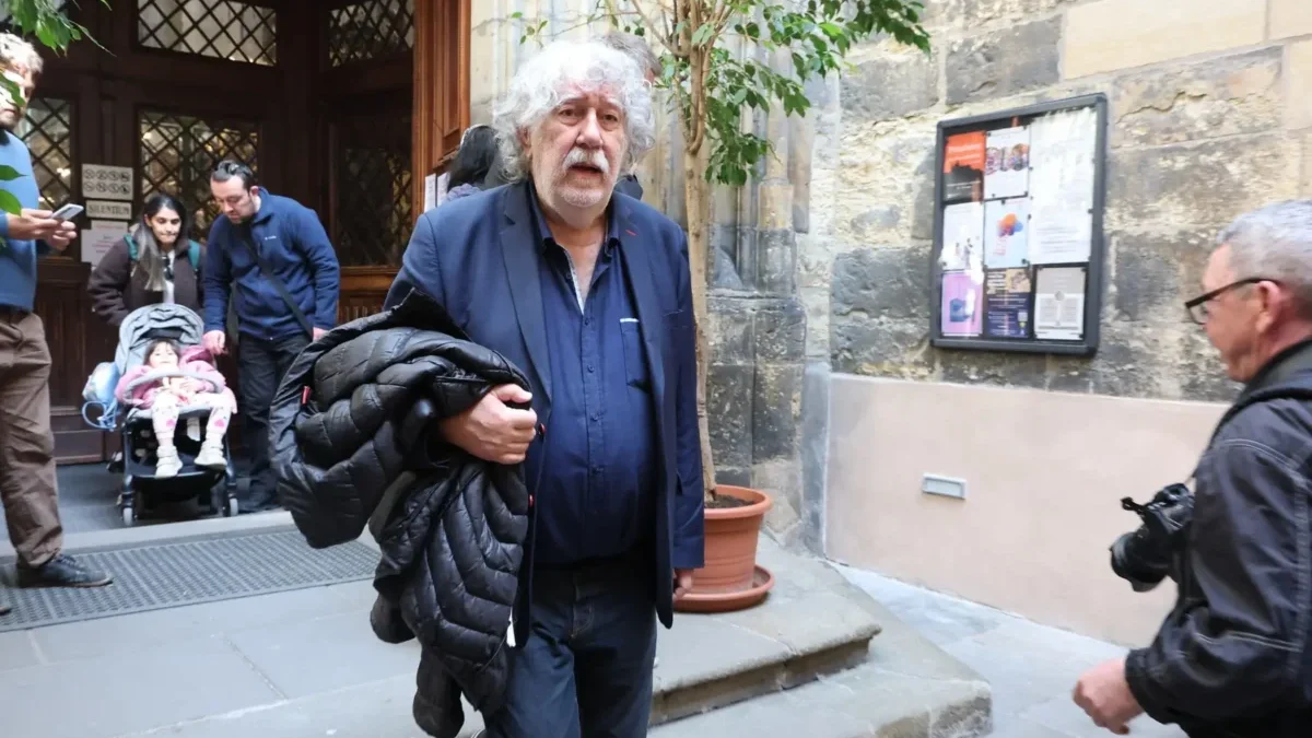 Older man with white hair and mustache in a blue blazer carrying a black puffer jacket on a city sidewalk.
