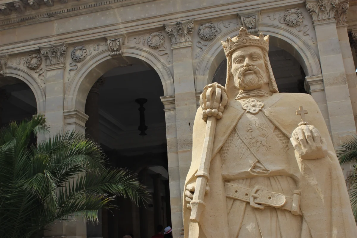 Stone statue of a crowned king in front of a historic arched façade with ornate carvings and columns.