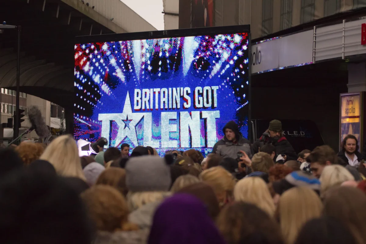 shutterstock_94503562 1 | TVGURU.cz Crowd gathered in an outdoor square watching a large LED screen showing'Britain's Got Talent'. The screen displays colorful graphics and text.