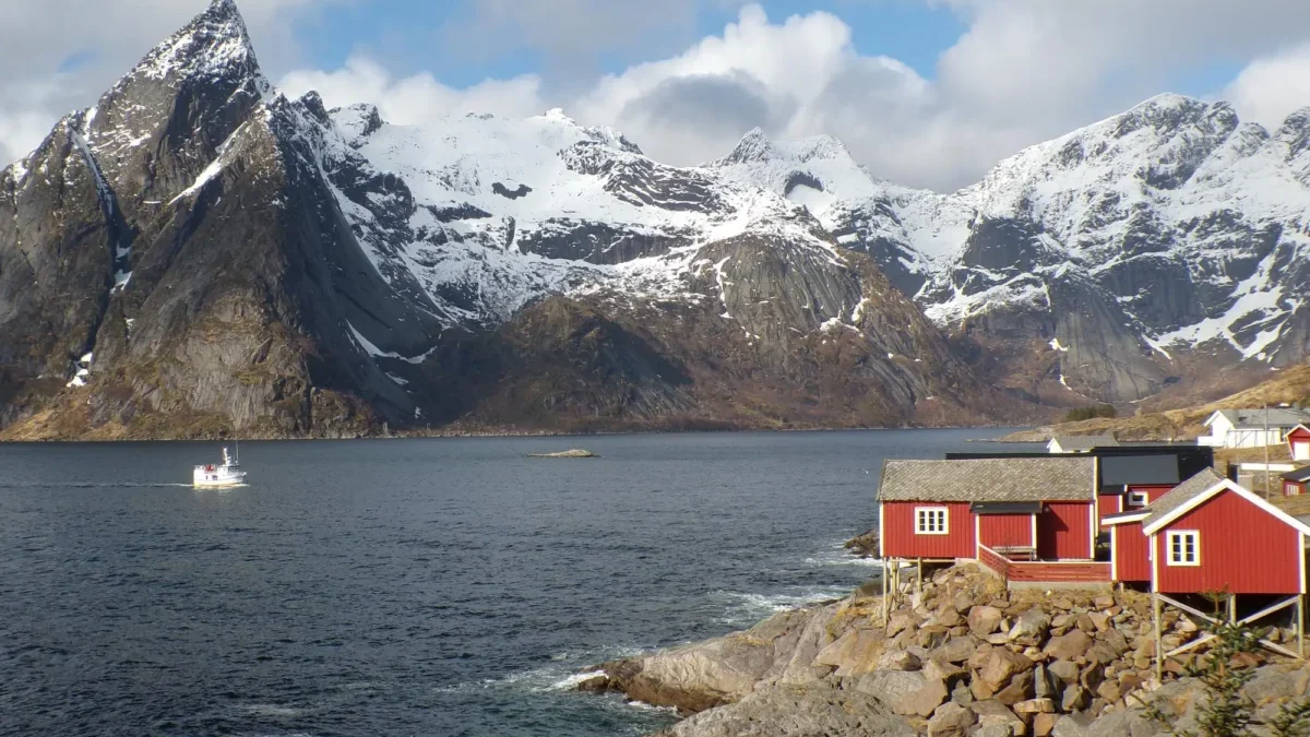 Snow-capped mountains meet a calm fjord, with red houses along a rocky shoreline and a small boat on the water.