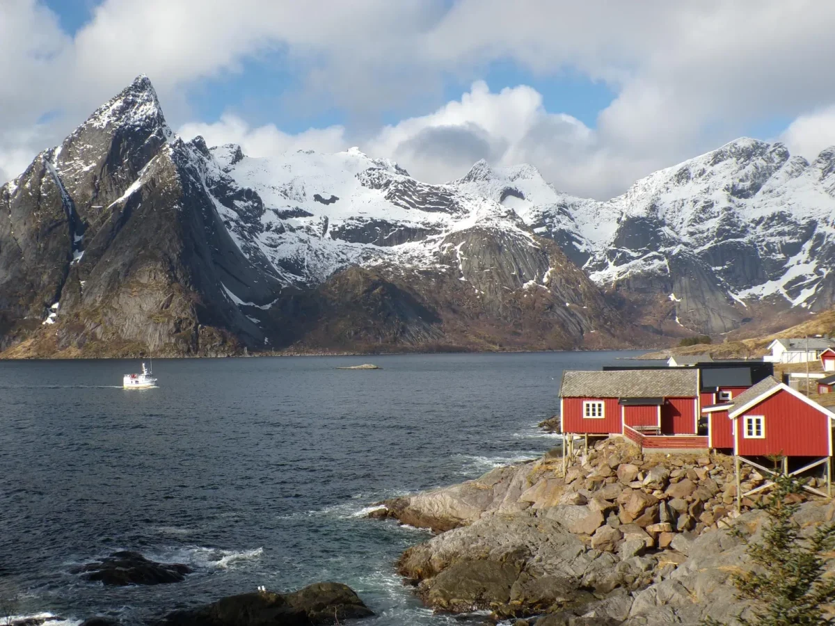 Snow-capped mountains meet a calm fjord, with red houses along a rocky shoreline and a small boat on the water.