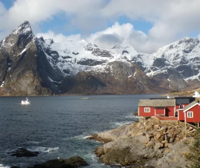 Snow-capped mountains meet a calm fjord, with red houses along a rocky shoreline and a small boat on the water.