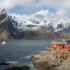 Snow-capped mountains meet a calm fjord, with red houses along a rocky shoreline and a small boat on the water.