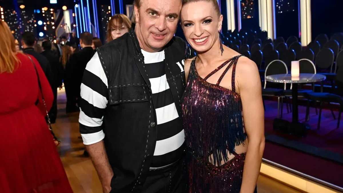 Two people pose for a photo at a festive event; man in black-and-white striped shirt and vest beside a smiling woman in a glittery dress, with blue stage lighting in the background.