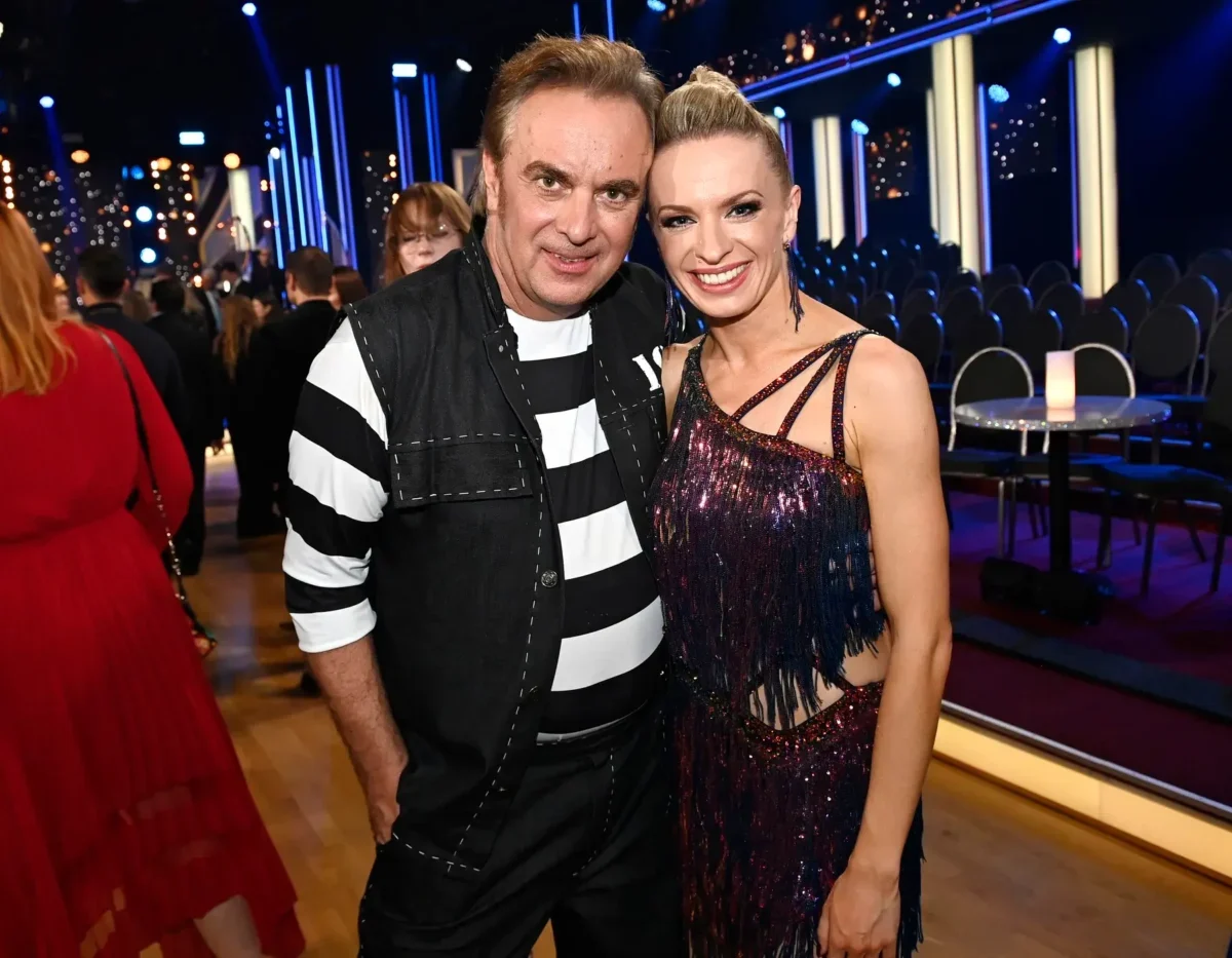 Two people pose for a photo at a festive event; man in black-and-white striped shirt and vest beside a smiling woman in a glittery dress, with blue stage lighting in the background.