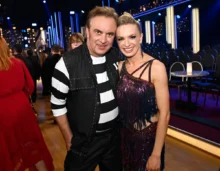 Two people pose for a photo at a festive event; man in black-and-white striped shirt and vest beside a smiling woman in a glittery dress, with blue stage lighting in the background.