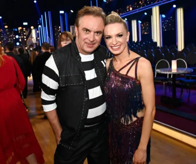 Two people pose for a photo at a festive event; man in black-and-white striped shirt and vest beside a smiling woman in a glittery dress, with blue stage lighting in the background.