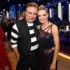 Two people pose for a photo at a festive event; man in black-and-white striped shirt and vest beside a smiling woman in a glittery dress, with blue stage lighting in the background.