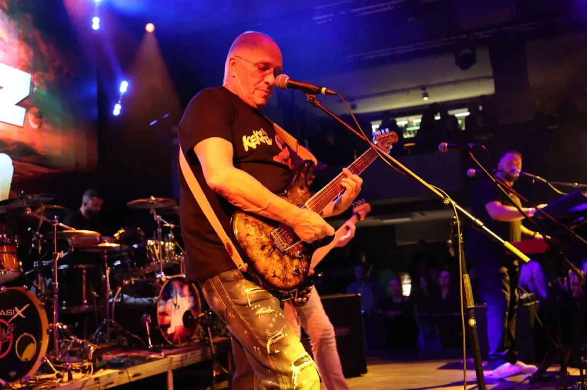 profimedia-1046623248 1 | TVGURU.cz Male guitarist on stage playing a marbled electric guitar, wearing a black t-shirt and ripped jeans, with a drummer in the background under colorful lights