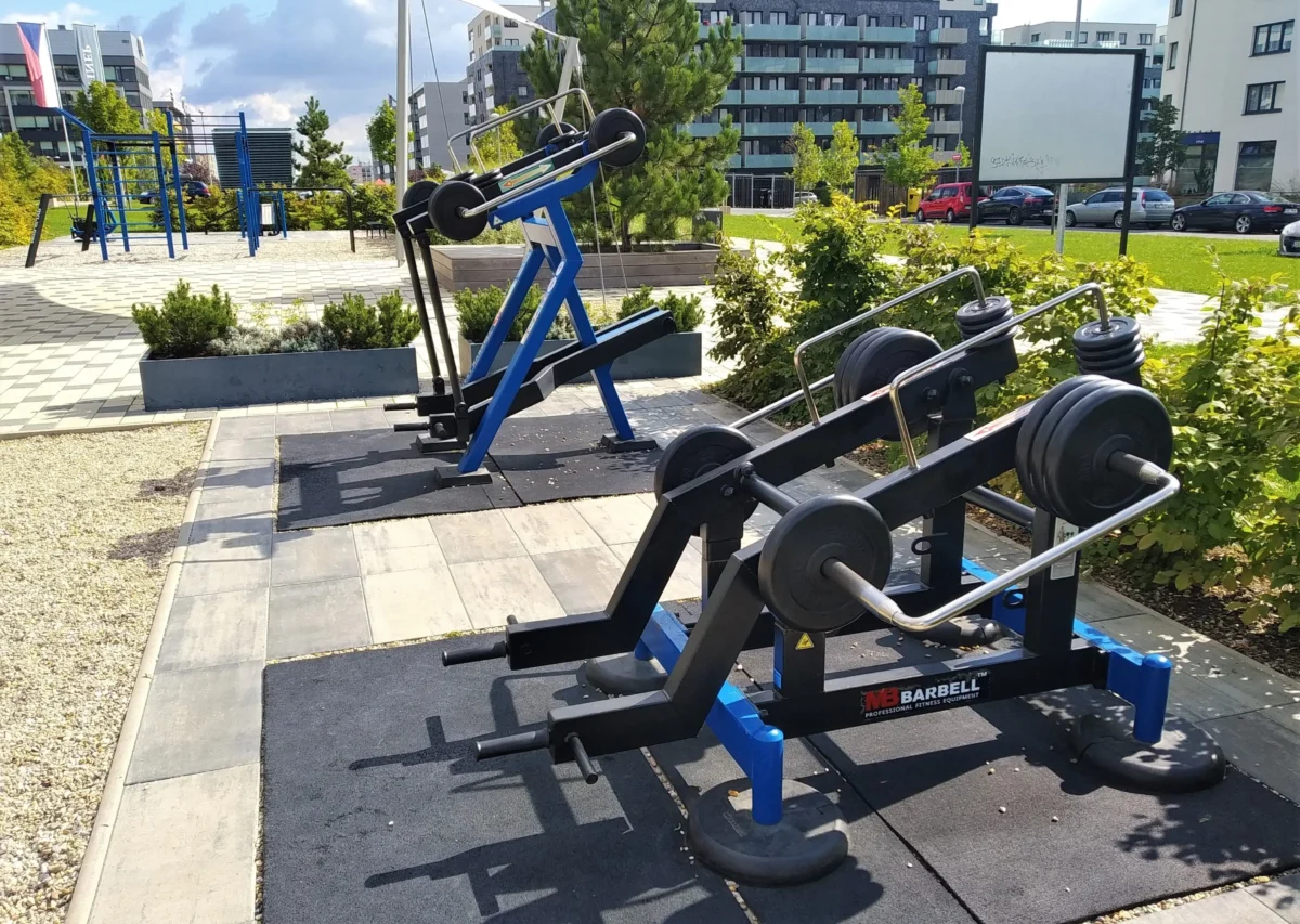 shutterstock_2207688619 1 | TVGURU.cz Outdoor gym area with blue weight machines and stacked barbells on a sunny day, with modern buildings in the background and green shrubs nearby.