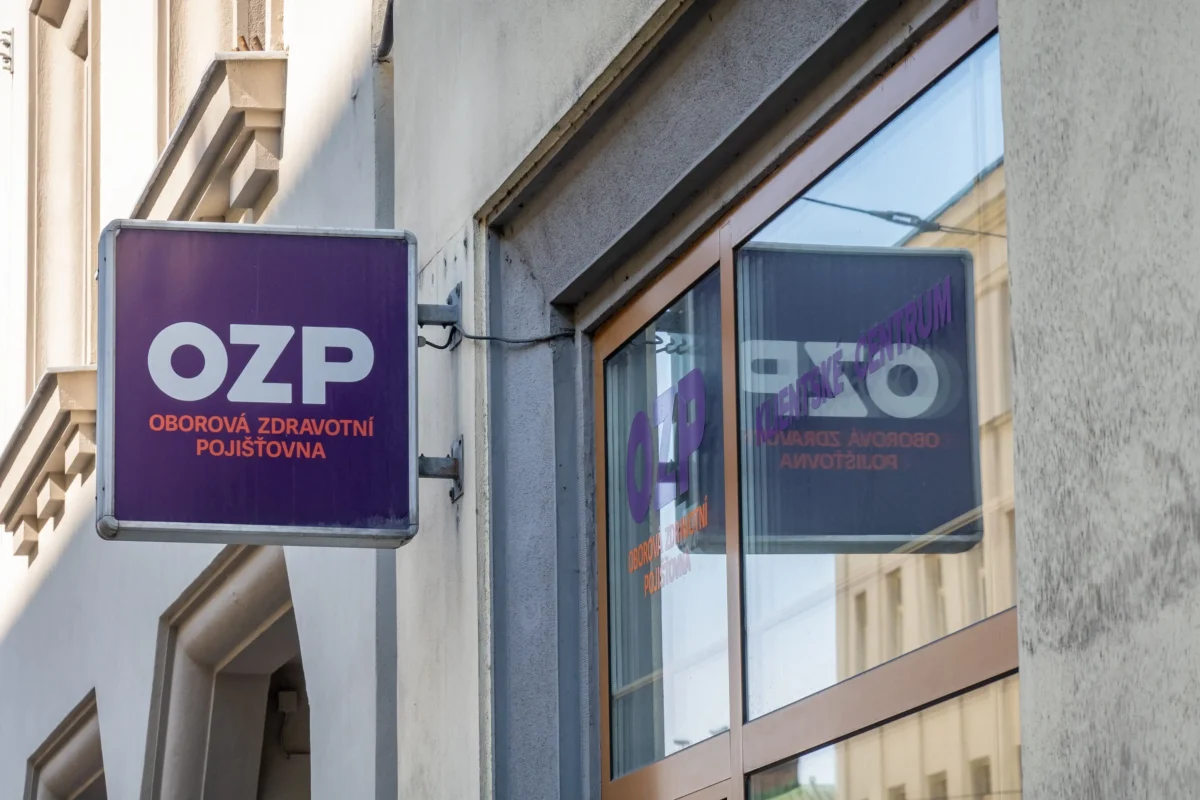 shutterstock_2488353463 1 | TVGURU.cz Purple OZP sign for Oborová zdravotní pojišťovna mounted on a beige building beside a glass storefront window.