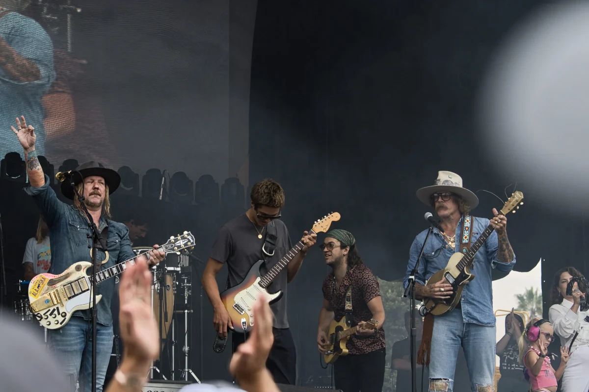 shutterstock_2720256875 1 | TVGURU.cz Band of musicians playing guitars on an outdoor stage, wearing denim and hats, with a cheering crowd in the foreground.