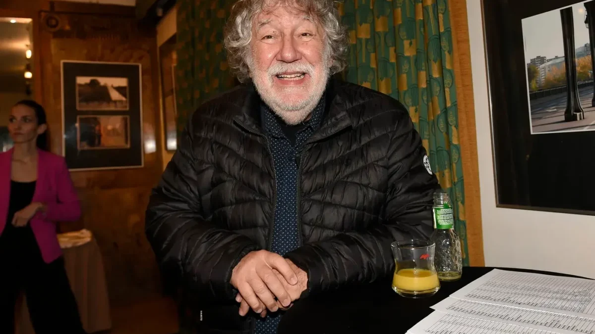 Smiling older man in a black puffer jacket leans on a round table with a glass of orange juice, a bottle, and papers at an indoor event.
