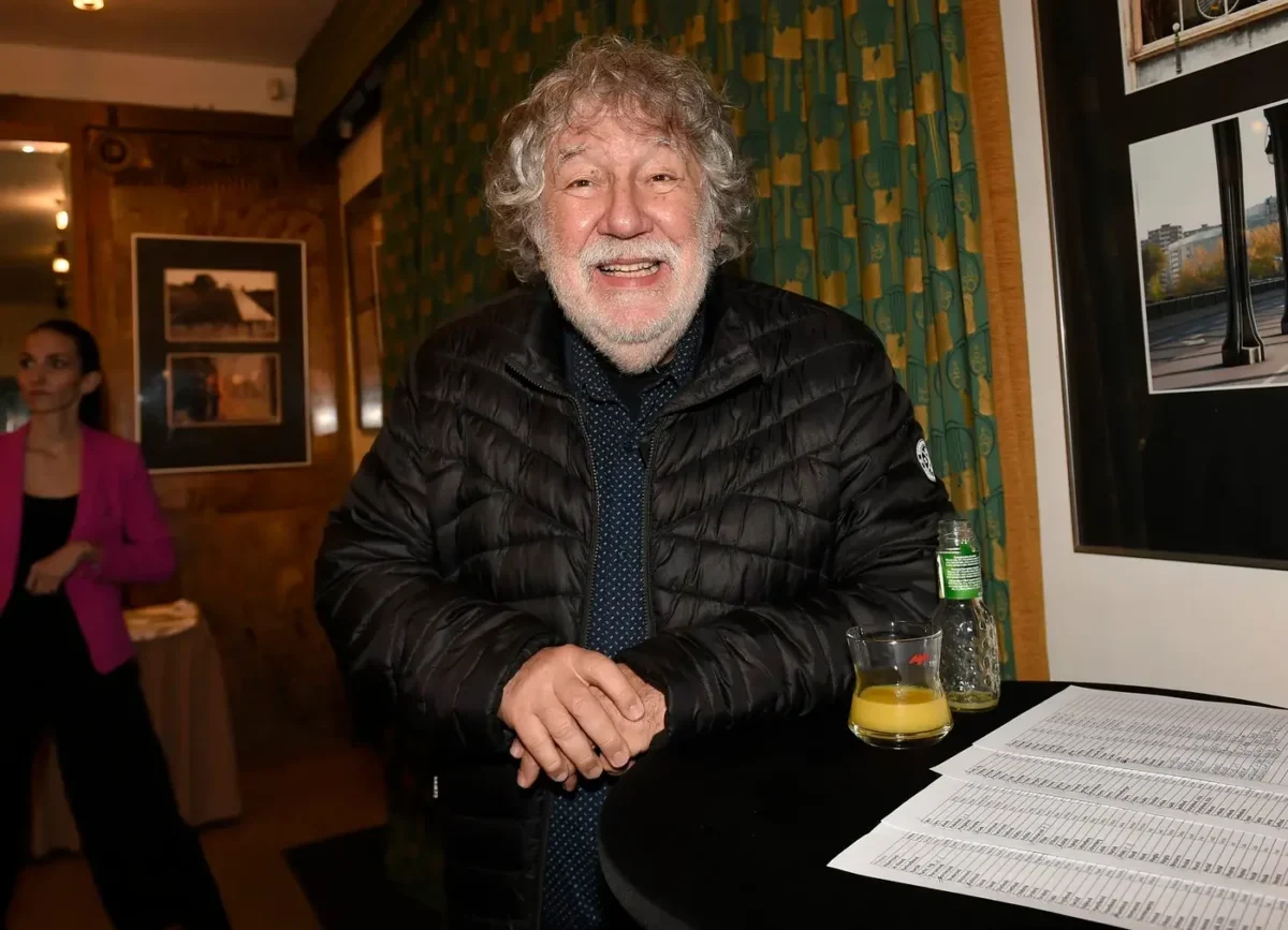Smiling older man in a black puffer jacket leans on a round table with a glass of orange juice, a bottle, and papers at an indoor event.