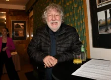 Smiling older man in a black puffer jacket leans on a round table with a glass of orange juice, a bottle, and papers at an indoor event.
