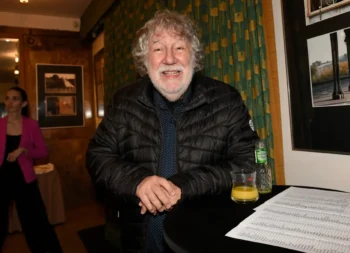 Smiling older man in a black puffer jacket leans on a round table with a glass of orange juice, a bottle, and papers at an indoor event.