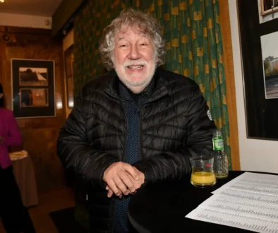 Smiling older man in a black puffer jacket leans on a round table with a glass of orange juice, a bottle, and papers at an indoor event.