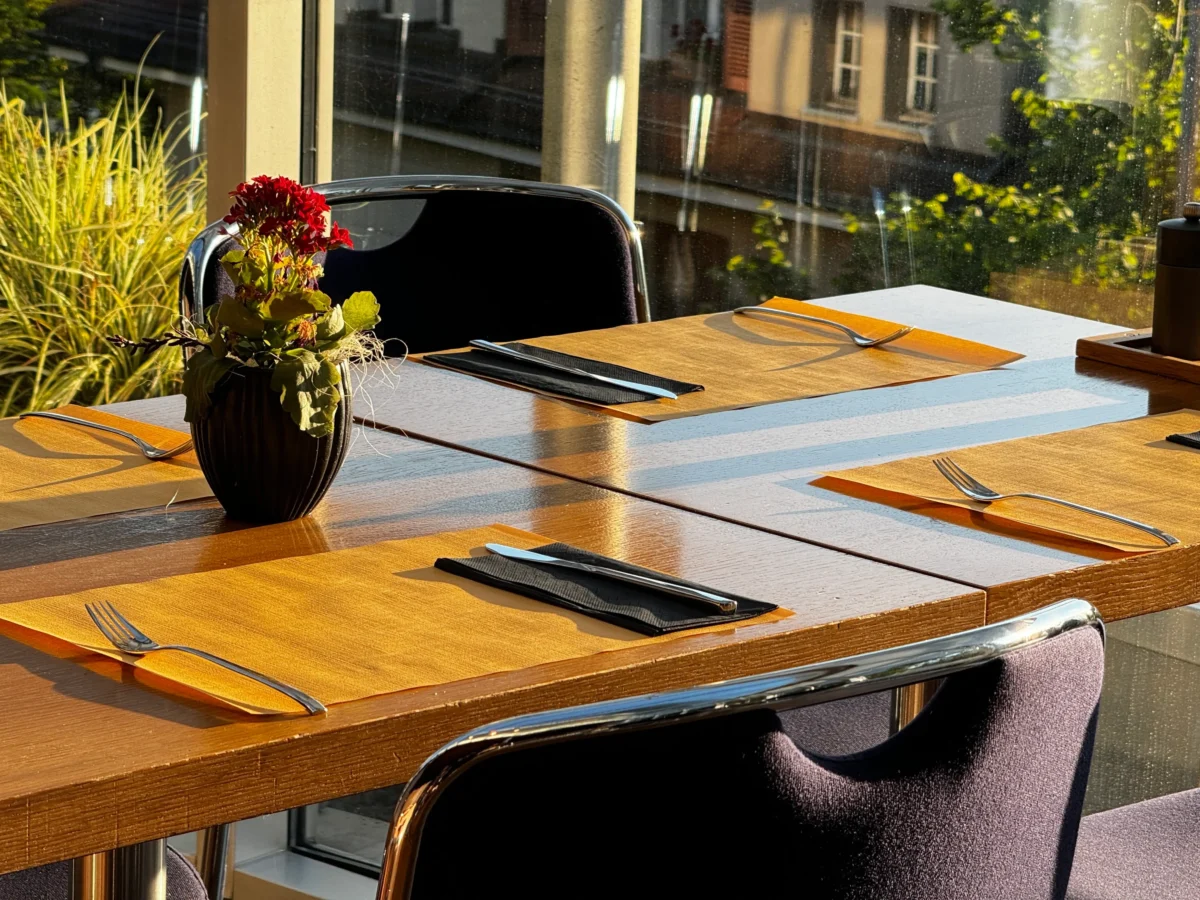 shutterstock_2740774573 1 | TVGURU.cz Sunlit cafe table set with orange placemats, black vase of red flowers, and silverware waiting for diners, through glass walls.