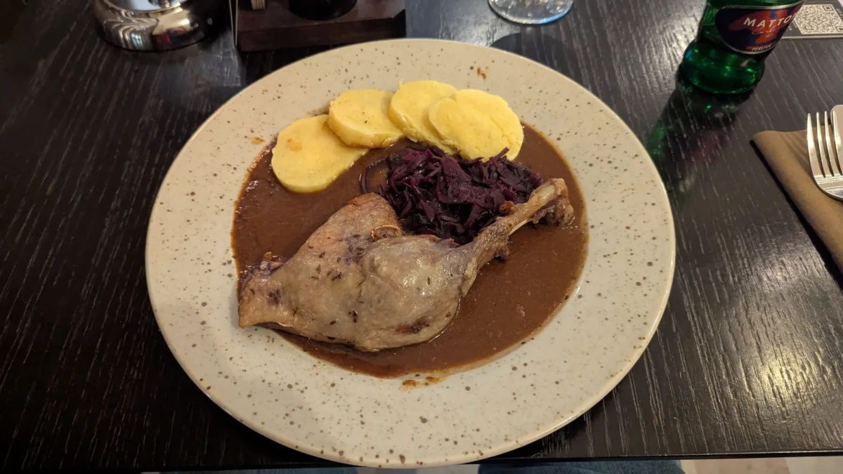 shutterstock_2775385733 1 | TVGURU.cz Braised meat in brown gravy with sliced plantains and sautéed red cabbage on a speckled plate at a dark wooden table.