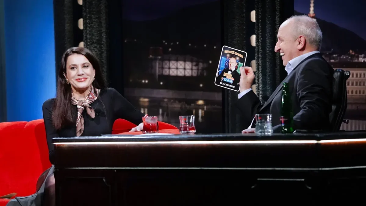 A woman and a man sit at a TV talk show desk, smiling; the man holds up a card featuring a photo and text. (Note: provides function/scene description without identifying individuals.)
