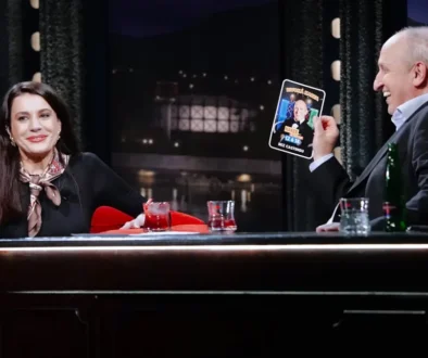 A woman and a man sit at a TV talk show desk, smiling; the man holds up a card featuring a photo and text. (Note: provides function/scene description without identifying individuals.)