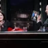 A woman and a man sit at a TV talk show desk, smiling; the man holds up a card featuring a photo and text. (Note: provides function/scene description without identifying individuals.)