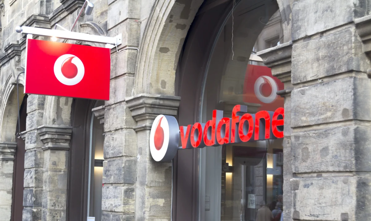 shutterstock_349718432 1 | TVGURU.cz Red Vodafone storefront sign on a historic stone building with a circular white logo and large red lettering in the window.