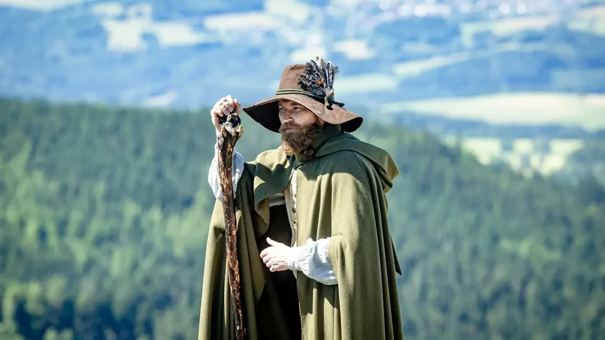 Muž v klobouku, s holí a s delšími vousy stojí venku a za ním jsou vidět rozlehlé lesy