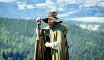 Muž v klobouku, s holí a s delšími vousy stojí venku a za ním jsou vidět rozlehlé lesy