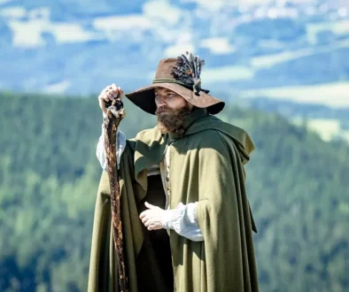 Muž v klobouku, s holí a s delšími vousy stojí venku a za ním jsou vidět rozlehlé lesy