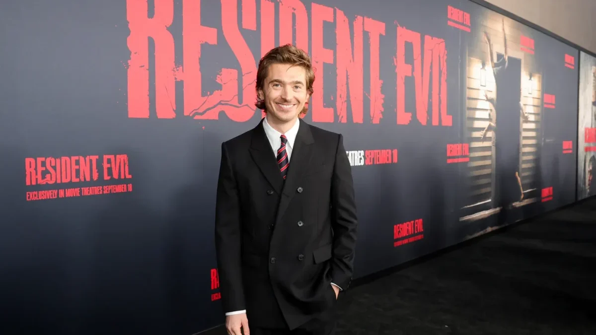 Smiling man in a black suit and red striped tie posing in front of a Resident Evil promotional wall at a premiere edge.