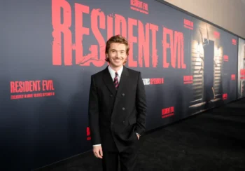 Smiling man in a black suit and red striped tie posing in front of a Resident Evil promotional wall at a premiere edge.
