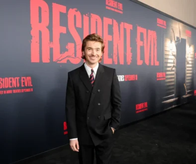 Smiling man in a black suit and red striped tie posing in front of a Resident Evil promotional wall at a premiere edge.