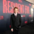 Smiling man in a black suit and red striped tie posing in front of a Resident Evil promotional wall at a premiere edge.