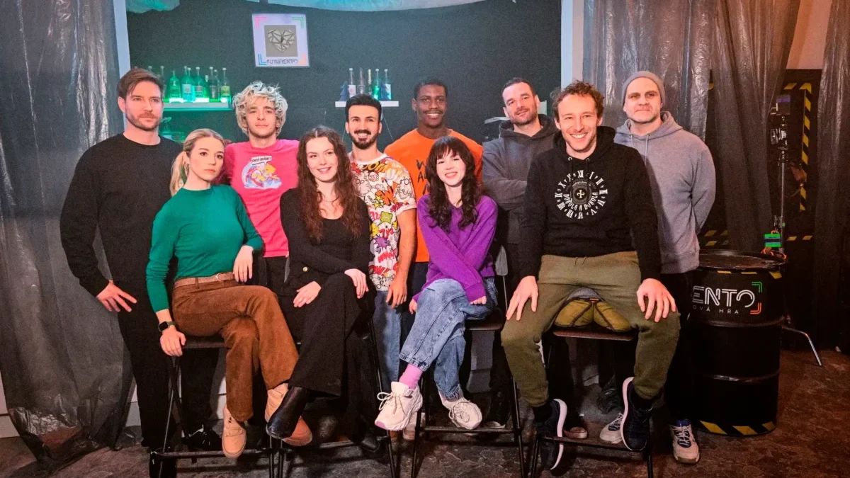 Twelve people posing together in a dim studio, some seated on stools, others standing in front of a bar-like backdrop with bottles and teal lighting.