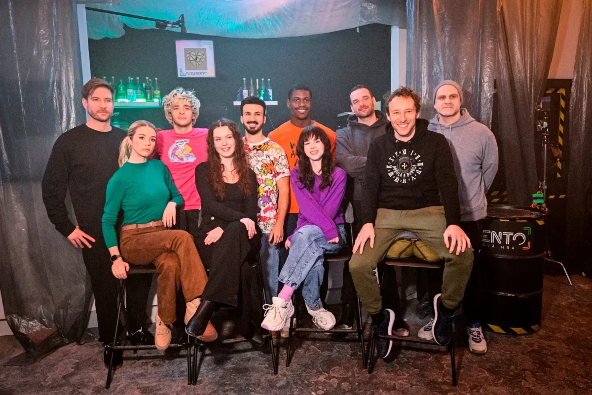Twelve people posing together in a dim studio, some seated on stools, others standing in front of a bar-like backdrop with bottles and teal lighting.