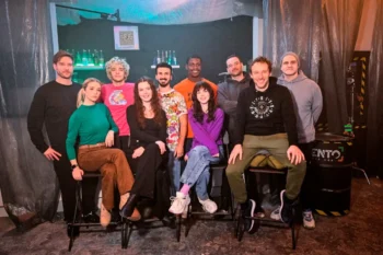 Twelve people posing together in a dim studio, some seated on stools, others standing in front of a bar-like backdrop with bottles and teal lighting.