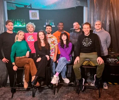 Twelve people posing together in a dim studio, some seated on stools, others standing in front of a bar-like backdrop with bottles and teal lighting.