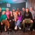 Twelve people posing together in a dim studio, some seated on stools, others standing in front of a bar-like backdrop with bottles and teal lighting.