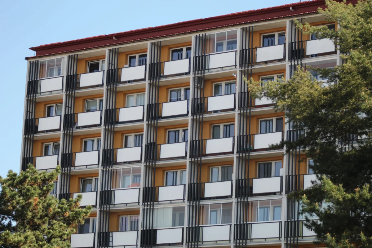 shutterstock_2619319857 1 | TVGURU.cz Modern apartment building with orange walls, white balconies, and black vertical railings against a clear blue sky; trees in foreground.