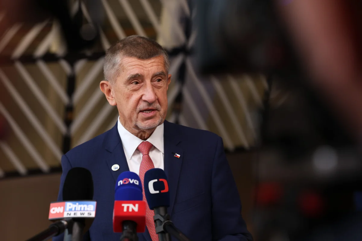 shutterstock_2814838687 1 | TVGURU.cz Man in a navy suit and red tie speaking at a press conference with multiple news microphones visible in front of him.