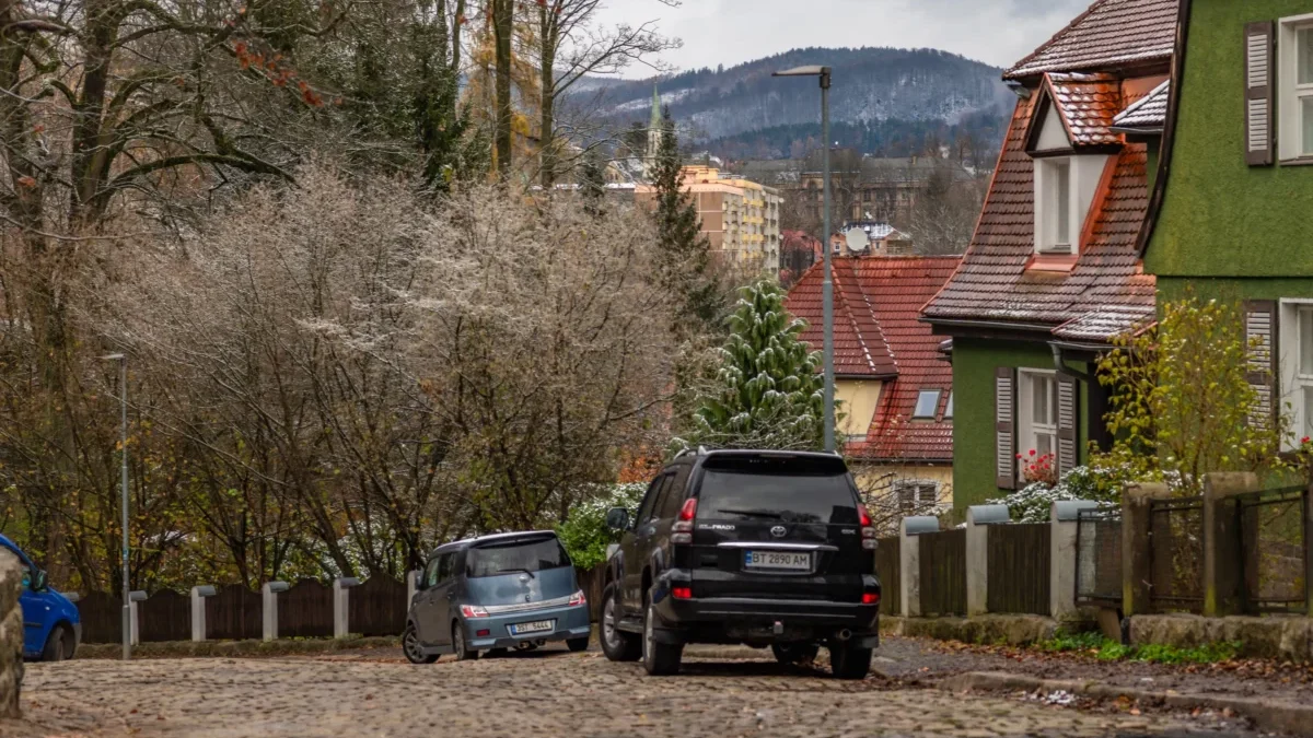 Wintery town street with cobblestones, parked cars, and houses with red-tiled roofs and a green exterior wall, set against snow-dusted hills.