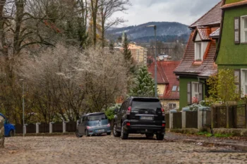 Wintery town street with cobblestones, parked cars, and houses with red-tiled roofs and a green exterior wall, set against snow-dusted hills.