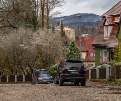 Wintery town street with cobblestones, parked cars, and houses with red-tiled roofs and a green exterior wall, set against snow-dusted hills.