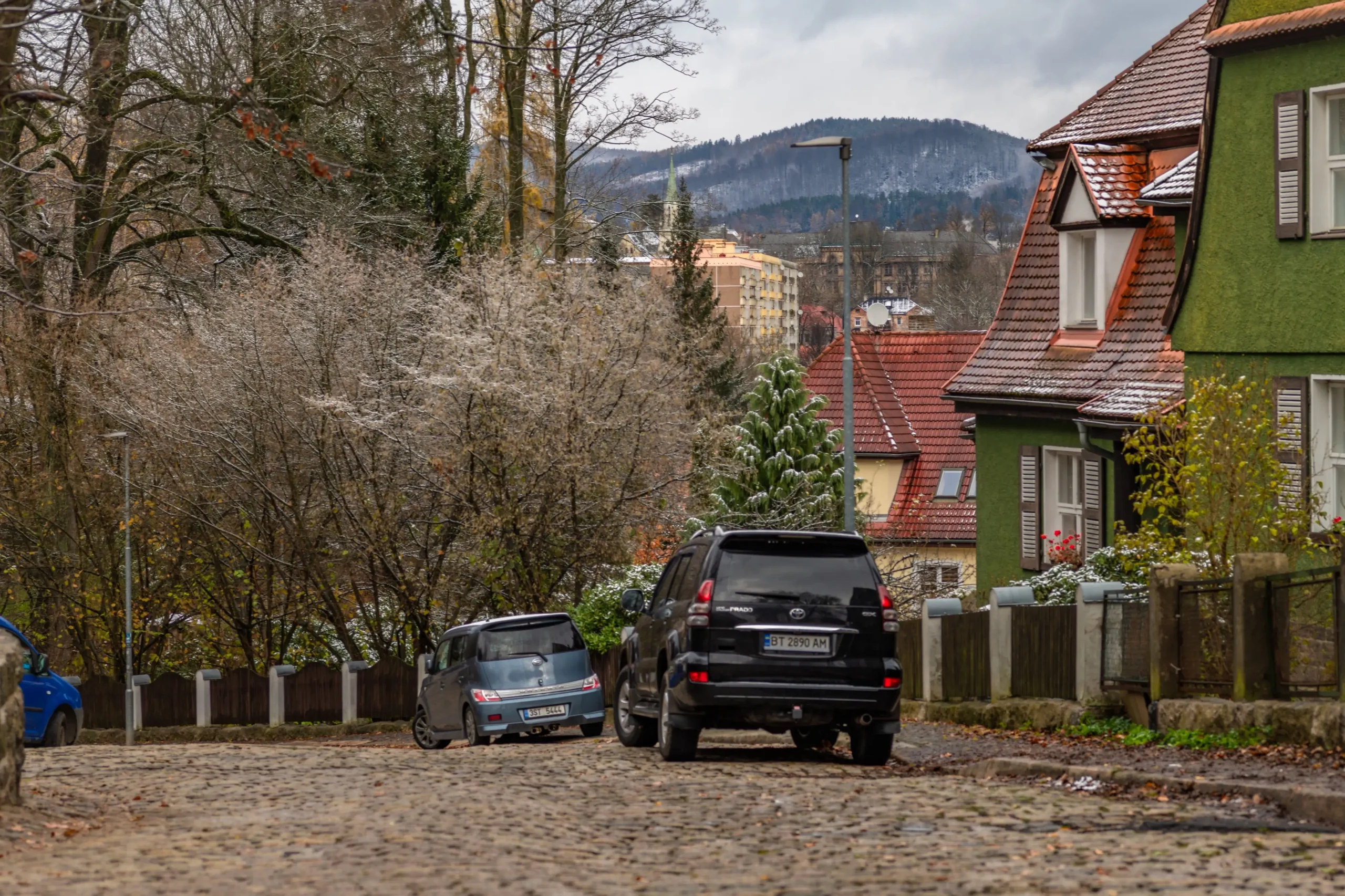 Wintery town street with cobblestones, parked cars, and houses with red-tiled roofs and a green exterior wall, set against snow-dusted hills.