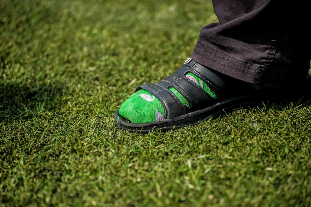 Close-up of a green socked foot in a black sandal resting on grass, with a dark pant leg nearby.
