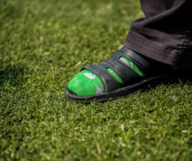 Close-up of a green socked foot in a black sandal resting on grass, with a dark pant leg nearby.
